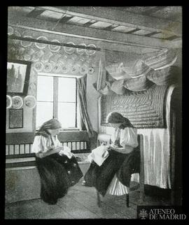 
[Dos mujeres cosiendo en el interior de una casa rural]
